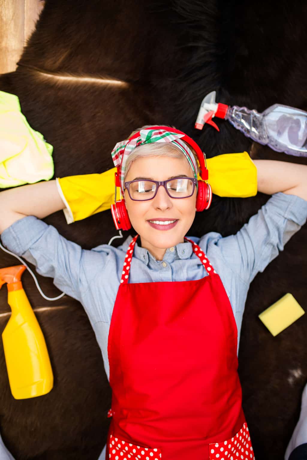 young woman laying down surrounded by cleaning supplies, eyes closed, with headphones on