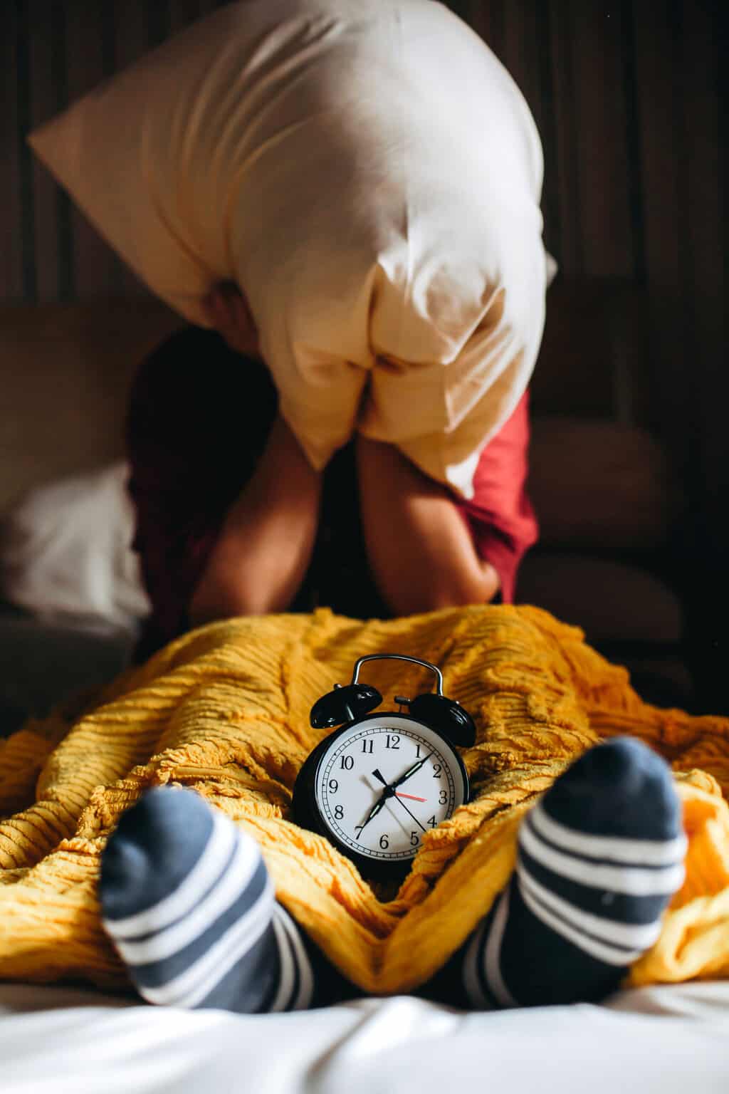 woman covering her head with a pillow, with old fashioned alarm clock on bed, sleep and mental health