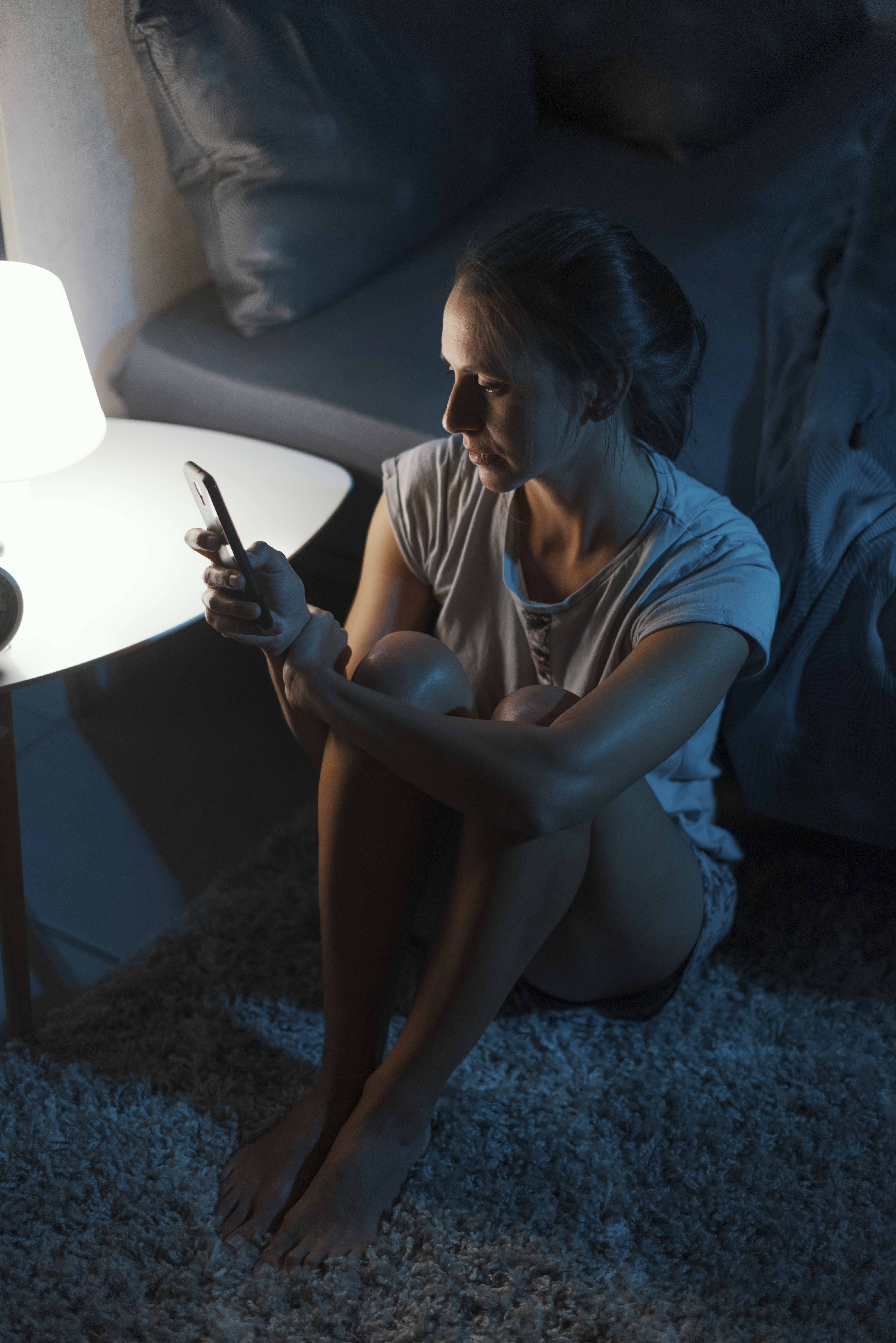 woman sitting on floor beside bed looking at her phone at night, sleep and mental health
